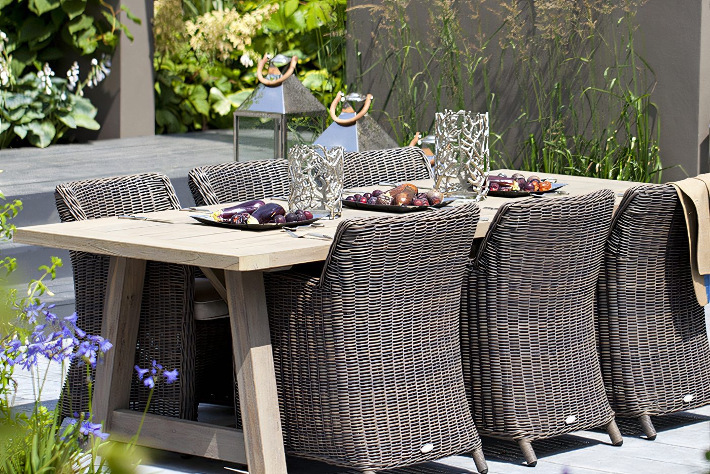 Une table et des chaises de rotin sur une terrasse de pavé, plantations autour, au soleil