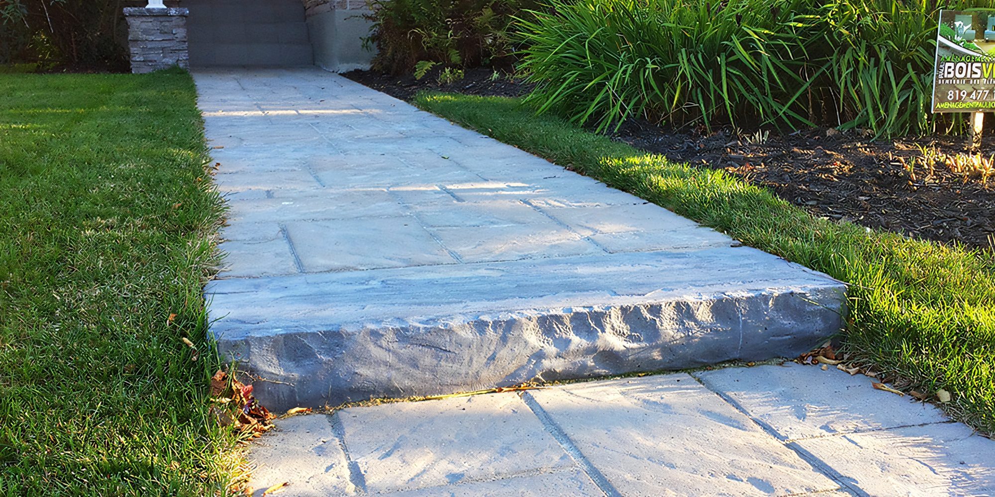 Trottoir de pavé de béton bordé de graminées, l’enseigne de chantier d’Aménagement Paul Boisvert plantée au fond